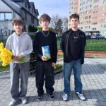 Three young men stand on a paved walkway outside a residential area, with a brick fence and apartment buildings in the background. The left person holds a bouquet of bright yellow daffodils; the middle person holds a blue gift box; the right person wears a black jacket and jeans.