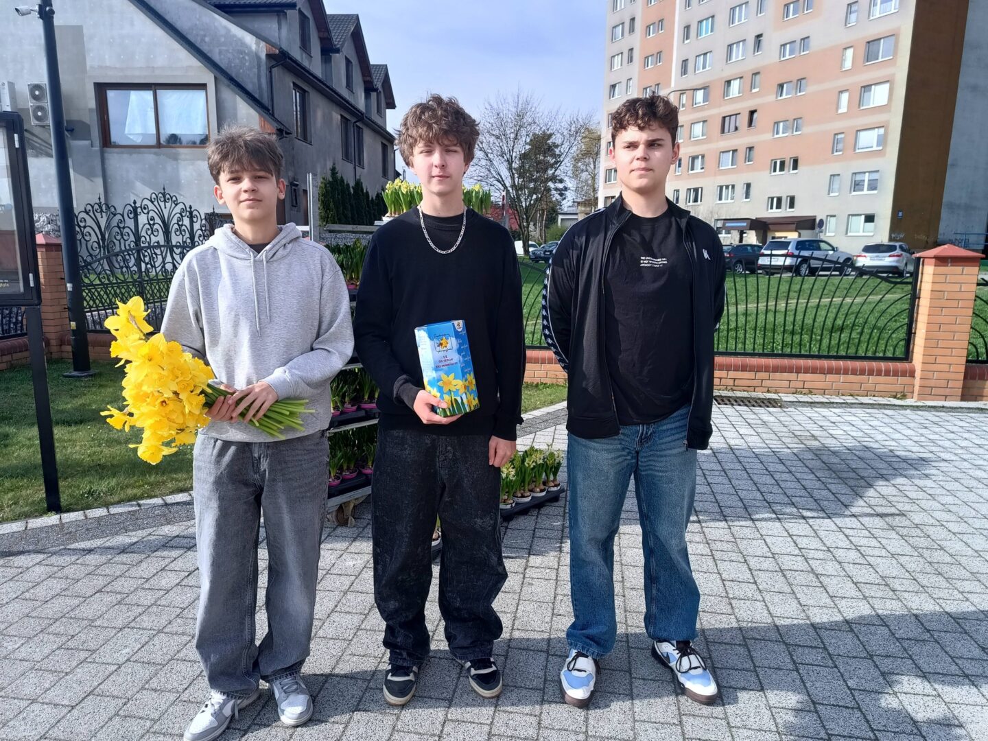 Three young men stand on a paved walkway outside a residential area, with a brick fence and apartment buildings in the background. The left person holds a bouquet of bright yellow daffodils; the middle person holds a blue gift box; the right person wears a black jacket and jeans.
