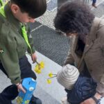A uniformed young person hands a yellow daffodil to a small child while an adult bends down to assist, on a paved outdoor area with other people in the background.