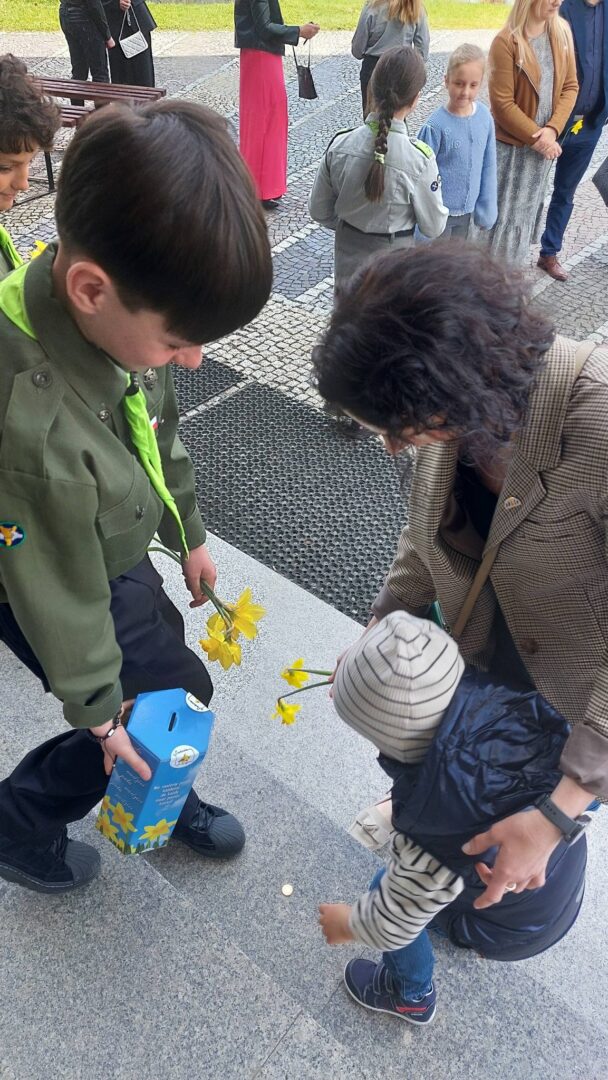 A uniformed young person hands a yellow daffodil to a small child while an adult bends down to assist, on a paved outdoor area with other people in the background.