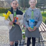 Two girls in scout uniforms stand on a cobblestone path; one holds yellow daffodils and a small card, the other a blue box with yellow flowers.