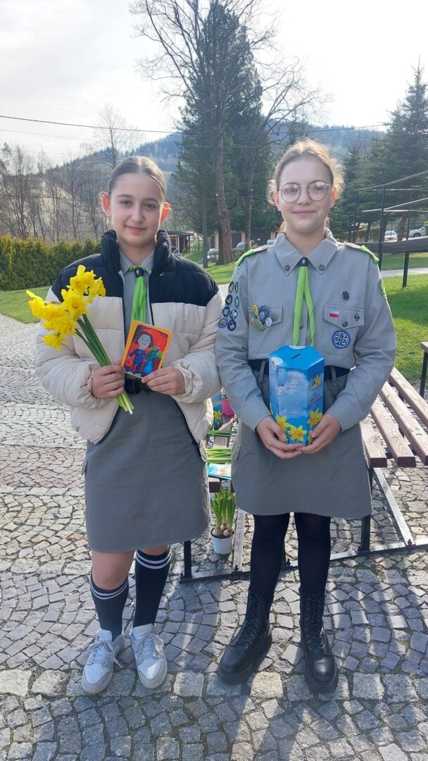 Two girls in scout uniforms stand on a cobblestone path; one holds yellow daffodils and a small card, the other a blue box with yellow flowers.