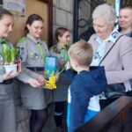 Group of Scouts in gray uniforms handing flowers and a book to a young boy and an older woman at a doorway outside a stone building.