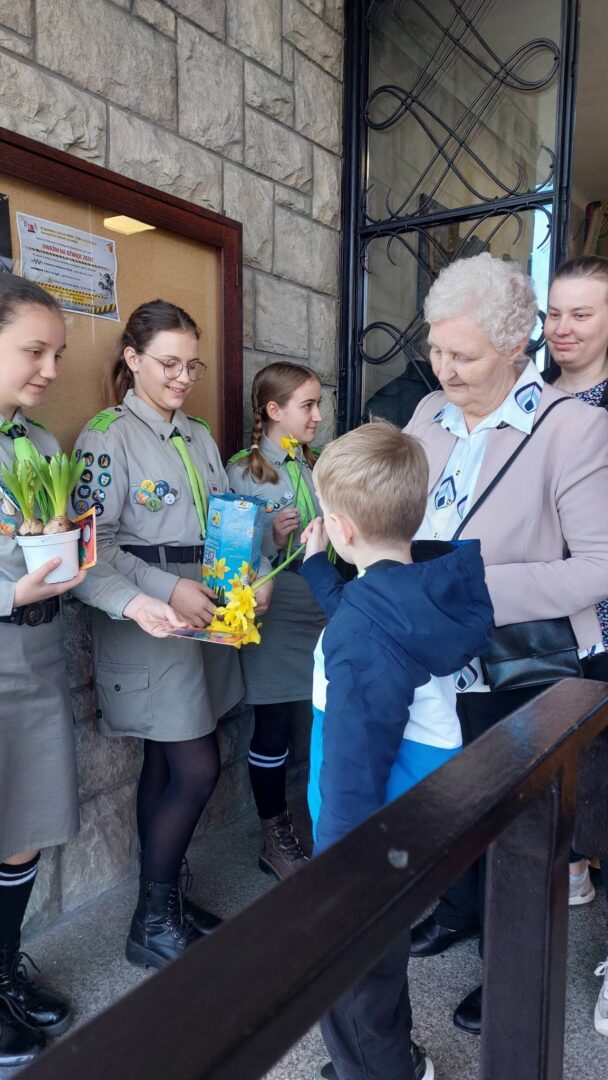 Group of Scouts in gray uniforms handing flowers and a book to a young boy and an older woman at a doorway outside a stone building.