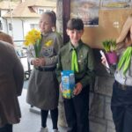 Three students in scout uniforms stand near a stone wall; left girl holds yellow daffodils, center boy holds a blue flower box, right girl holds a potted plant.