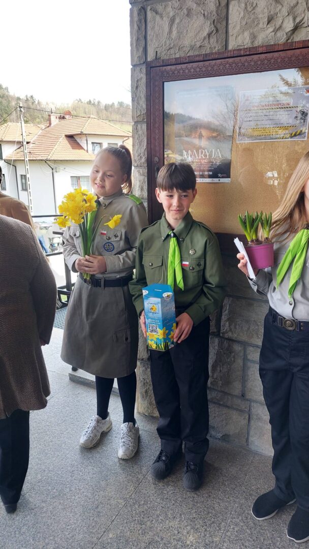 Three students in scout uniforms stand near a stone wall; left girl holds yellow daffodils, center boy holds a blue flower box, right girl holds a potted plant.