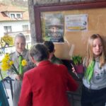 Children in gray uniforms and green scarves hand yellow daffodils to a person in a red coat near a stone wall with a bulletin board and posters outside a building.