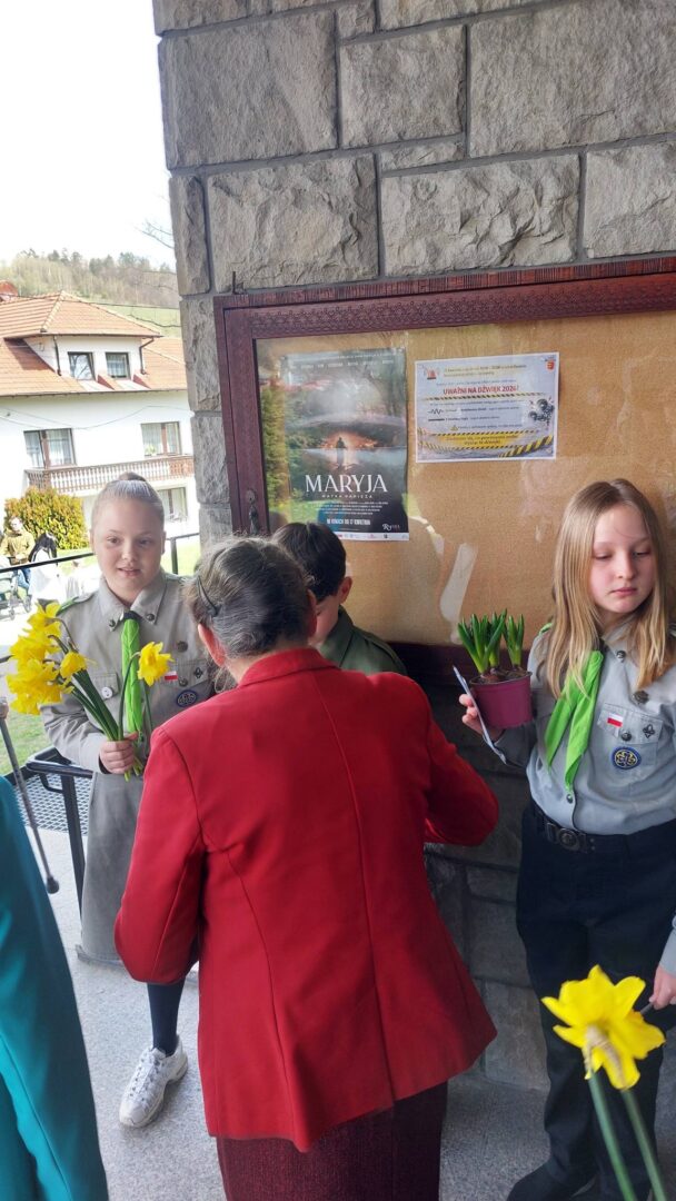 Children in gray uniforms and green scarves hand yellow daffodils to a person in a red coat near a stone wall with a bulletin board and posters outside a building.