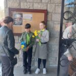 Two children in uniform stand by a bulletin board on a stone wall; the boy holds a blue gift bag and the girl, a bouquet of yellow daffodils, as a woman approaches nearby.