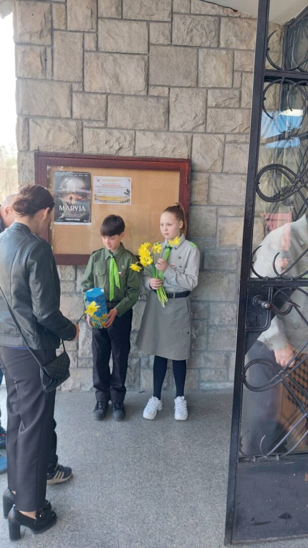 Two children in uniform stand by a bulletin board on a stone wall; the boy holds a blue gift bag and the girl, a bouquet of yellow daffodils, as a woman approaches nearby.