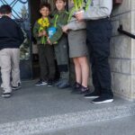 Group of children in uniform standing on school steps outside an entrance, some holding yellow flowers and a blue book, likely after a ceremony or event.