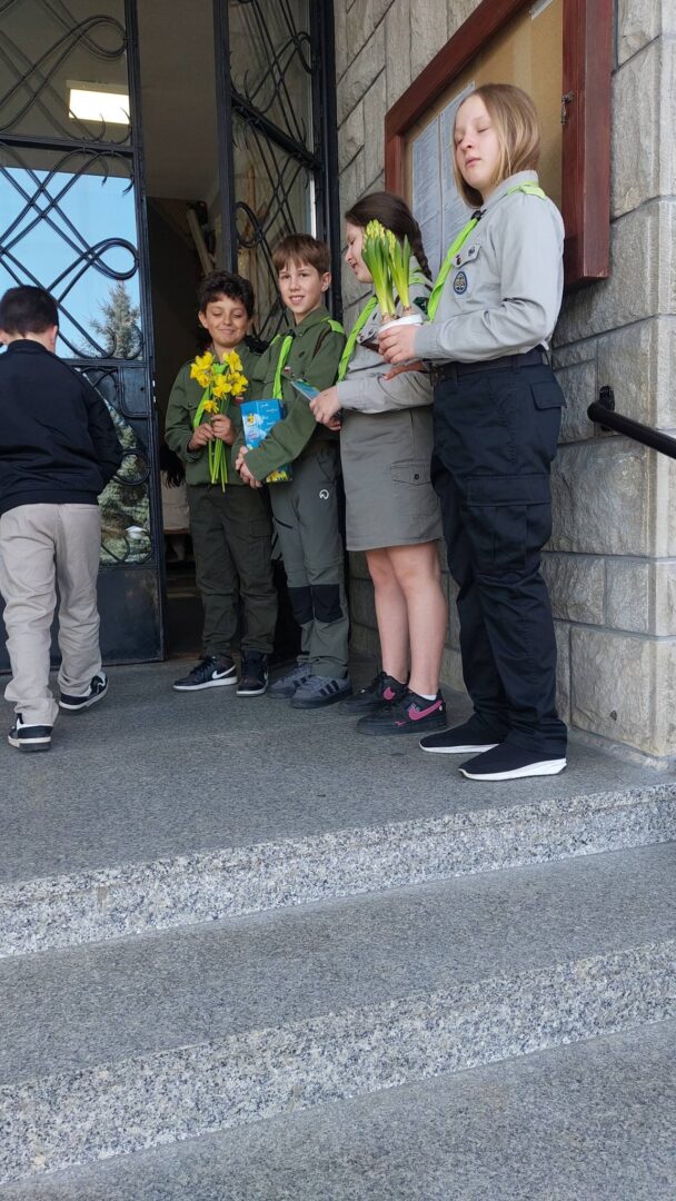 Group of children in uniform standing on school steps outside an entrance, some holding yellow flowers and a blue book, likely after a ceremony or event.