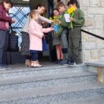 Children and adults on stone steps outside a building exchange yellow daffodils and papers.