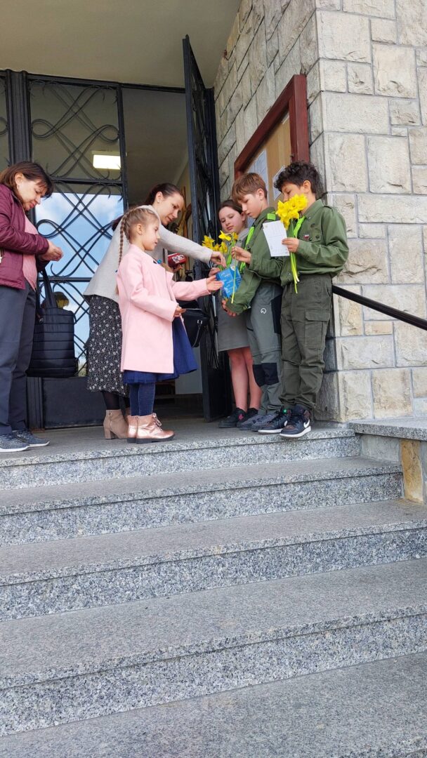Children and adults on stone steps outside a building exchange yellow daffodils and papers.