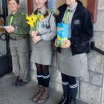 Three young scouts in uniform stand outside a stone building; center girl holds yellow daffodils, right girl holds a blue booklet.