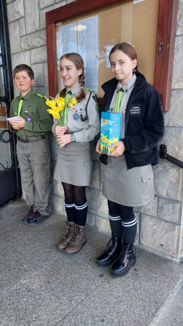 Three young scouts in uniform stand outside a stone building; center girl holds yellow daffodils, right girl holds a blue booklet.