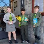 Three children in scout-style uniforms stand outside a stone building, the girl holding a pot of yellow flowers while the two boys hold flowers and cards/gift items.