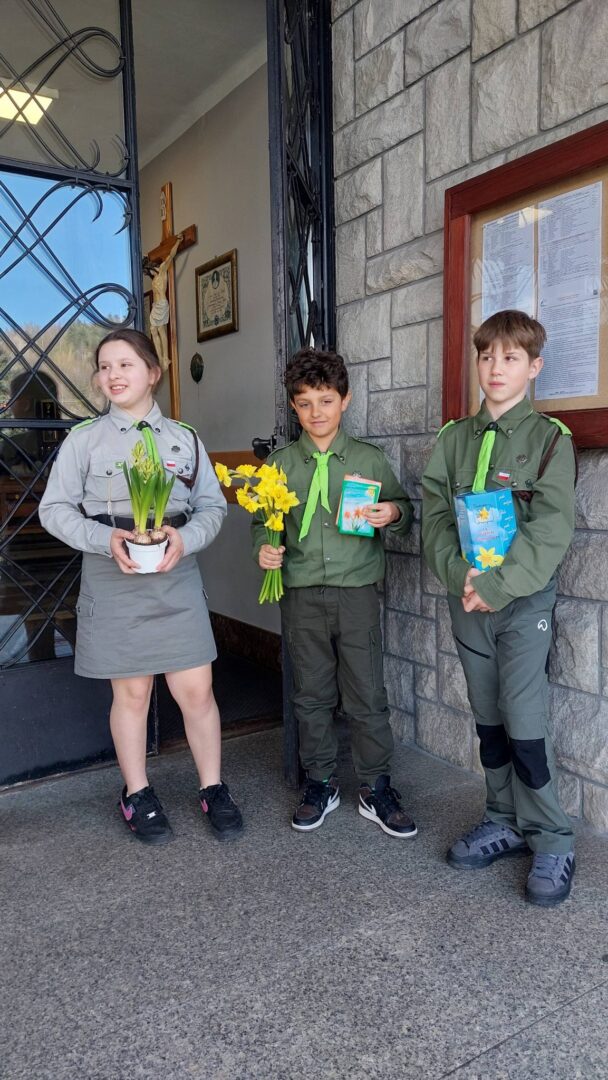 Three children in scout-style uniforms stand outside a stone building, the girl holding a pot of yellow flowers while the two boys hold flowers and cards/gift items.