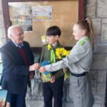 Older man in a suit shakes hands with a young boy in a green scout uniform, while a girl in a gray uniform hands him yellow flowers and a gift.