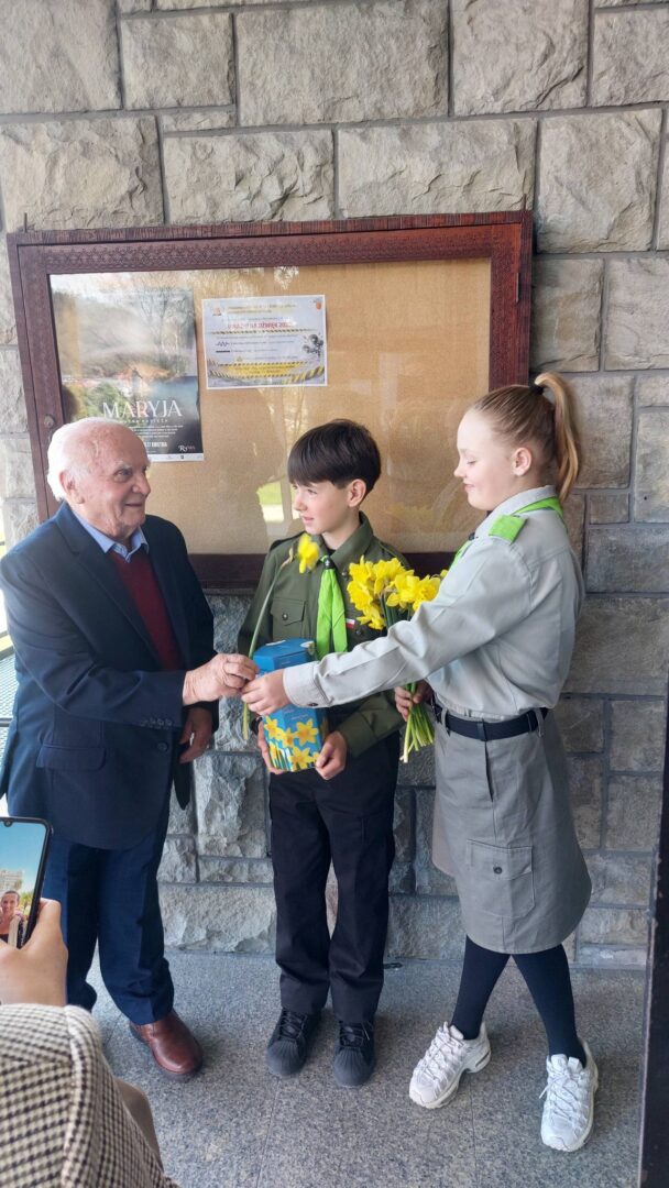 Older man in a suit shakes hands with a young boy in a green scout uniform, while a girl in a gray uniform hands him yellow flowers and a gift.