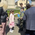 Young girl in a pink dress hands a certificate to a boy in a green uniform while adults look on outdoors near a building and bare trees.