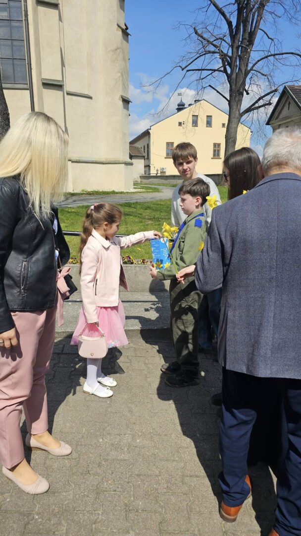 Girl in a pink dress hands a blue certificate to a boy in a green uniform as adults watch outdoors near a pale building at a ceremony.