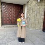 Woman in a beige coat standing outside a building, holding a bouquet of yellow daffodils and a blue book.