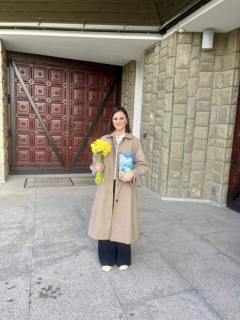 Woman in a beige coat standing outside a building, holding a bouquet of yellow daffodils and a blue book.