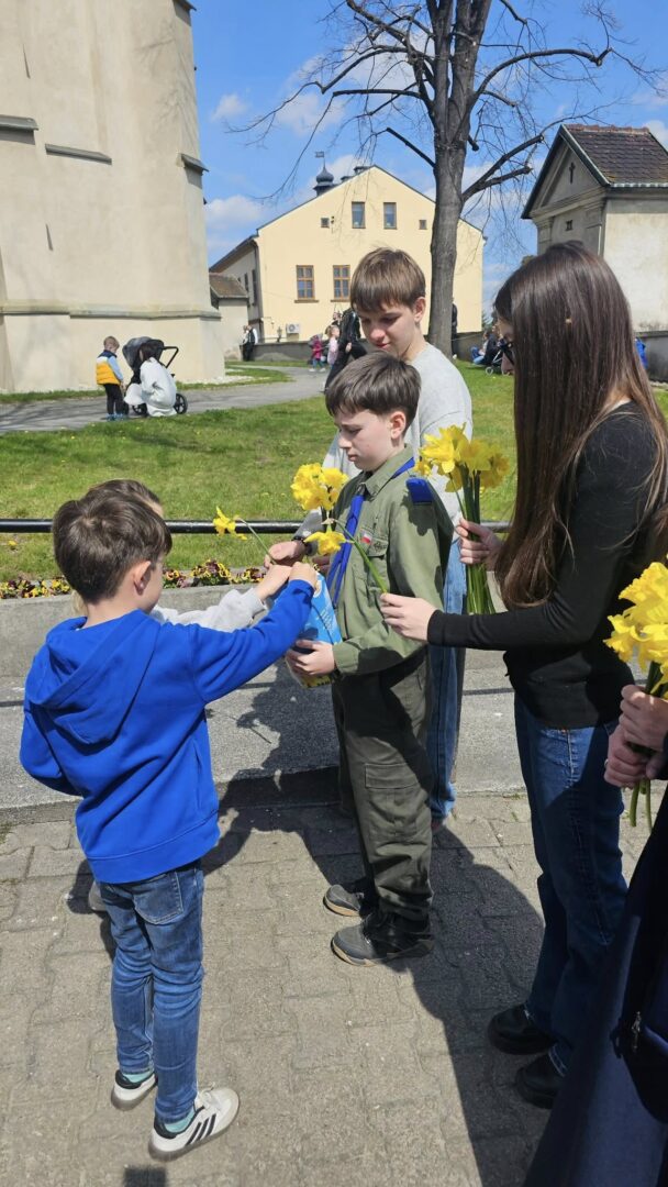 Child in a blue hoodie hands a yellow daffodil to a younger boy in olive overalls while others hold flowers nearby outside a campus-like area on a sunny day.