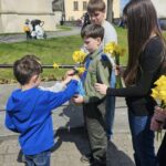 Group of children outdoors on a sunny day, as a boy in green overalls receives a yellow daffodil from a younger friend while others hold more flowers.
