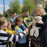 Group of schoolchildren presenting yellow daffodils to a smiling woman holding a baby outdoors.