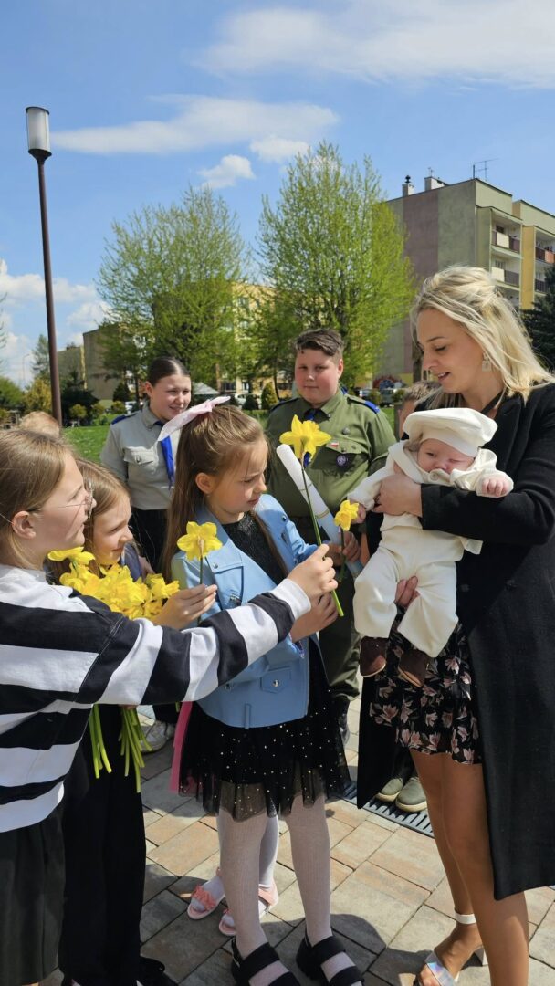 Group of schoolchildren presenting yellow daffodils to a smiling woman holding a baby outdoors.