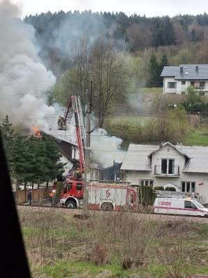 House in a suburban area on fire with heavy smoke, firefighters on a ladder truck directing water toward the flames.