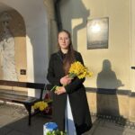 Young woman in a dark coat holding a bouquet of yellow flowers outside a building with a plaque on the wall.