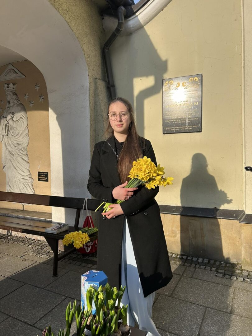 Young woman in a dark coat holding a bouquet of yellow flowers outside a building with a plaque on the wall.