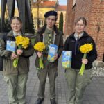 Three young students in green uniforms stand on a paved path outdoors, each holding a bouquet of yellow daffodils and a decorative blue box.