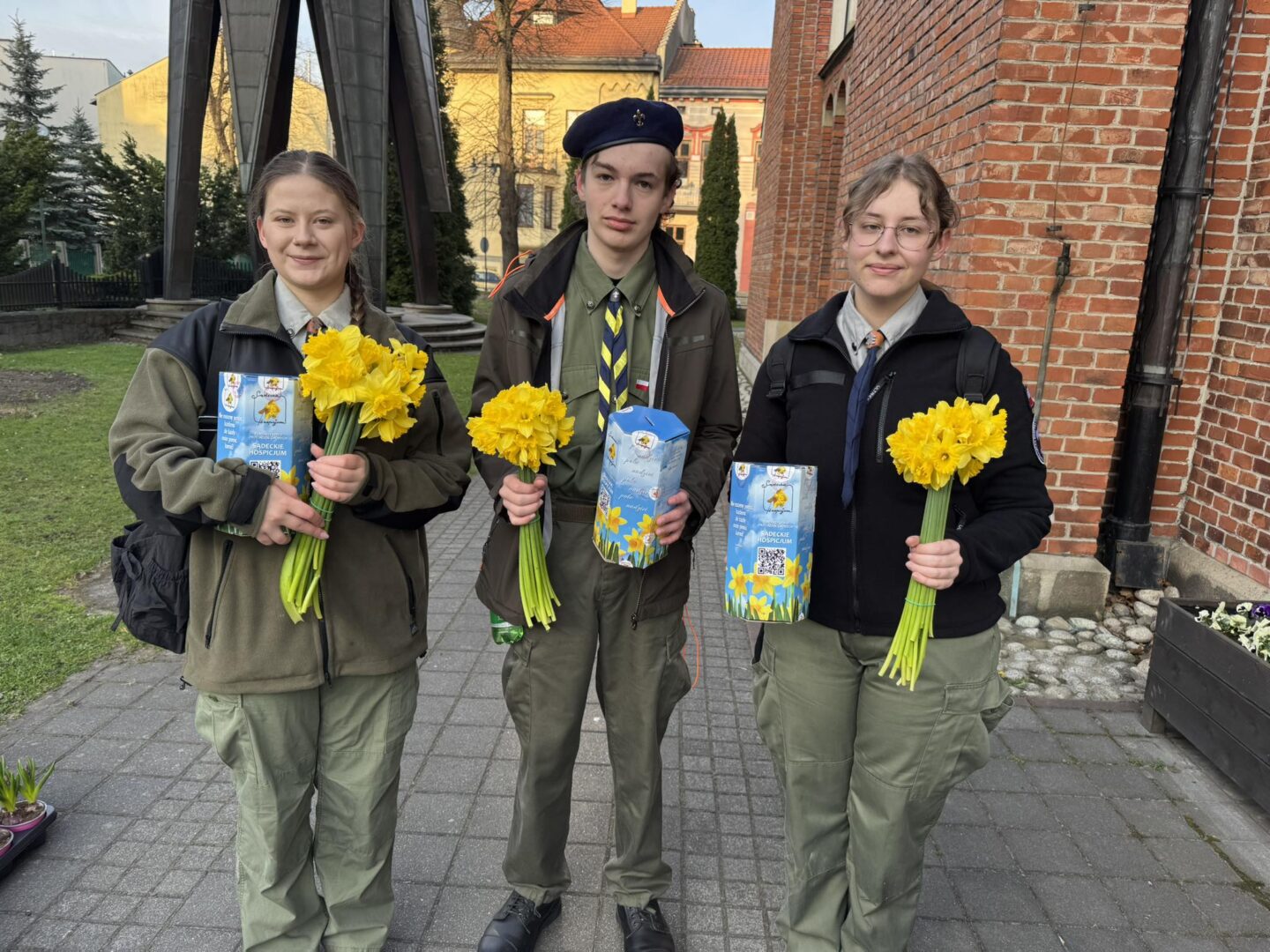 Three young students in green uniforms stand on a paved path outdoors, each holding a bouquet of yellow daffodils and a decorative blue box.