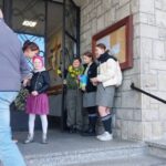 Group of children in school uniforms standing on church steps near an open door, some holding flowers and a book.