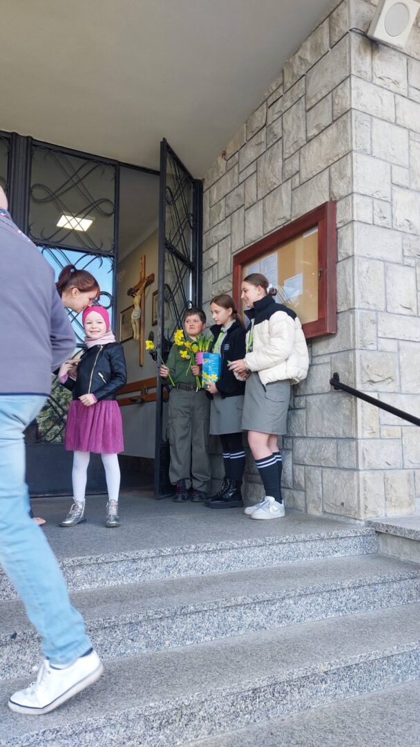Group of children in school uniforms standing on church steps near an open door, some holding flowers and a book.