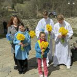 Five people outdoors—two altar servers in white robes and three civilians—holding bright yellow daffodil bouquets.