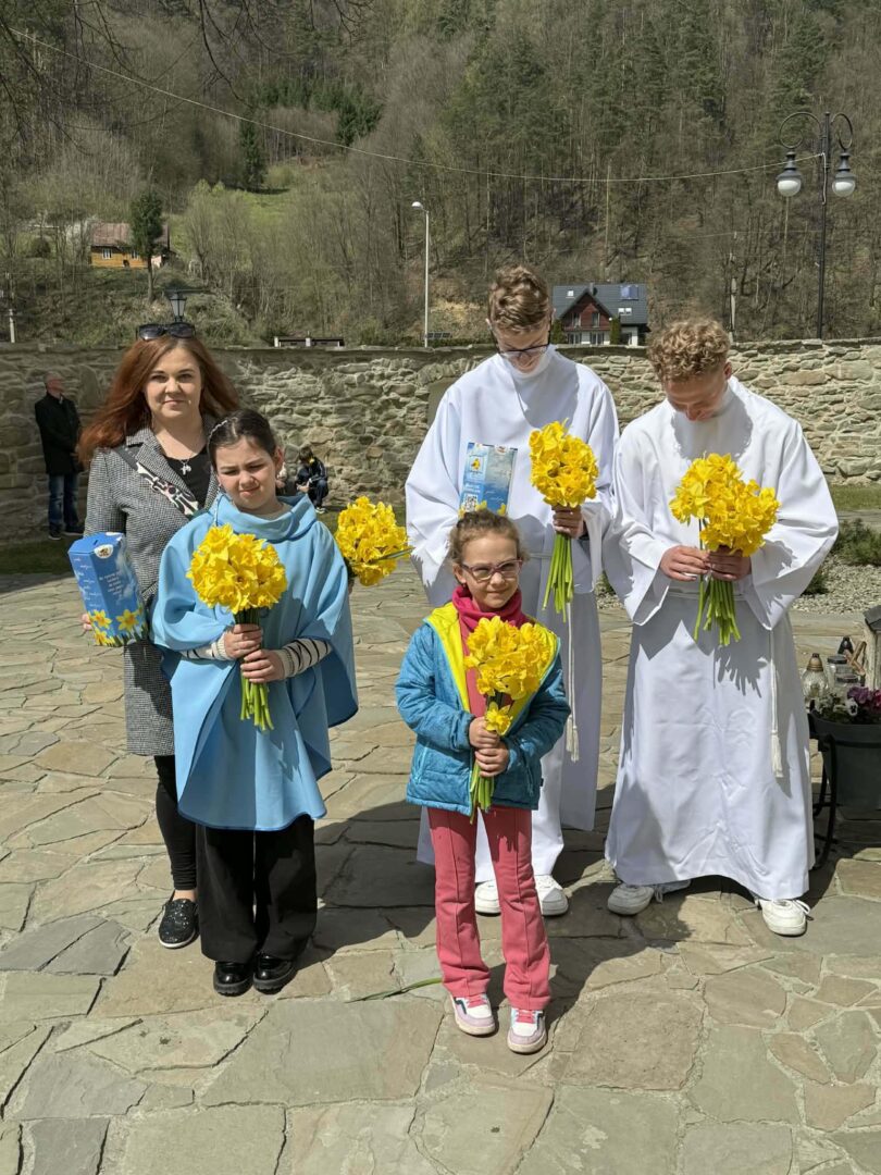 Five people outdoors—two altar servers in white robes and three civilians—holding bright yellow daffodil bouquets.