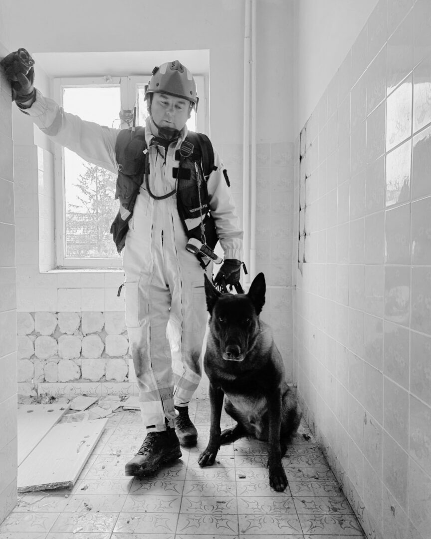 Firefighter in protective gear with a working dog inside a dilapidated tiled room, both looking forward.