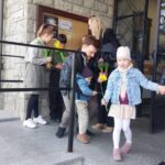Children holding daffodils and lining up at a stone building entrance; a girl in a denim jacket stands in the foreground.