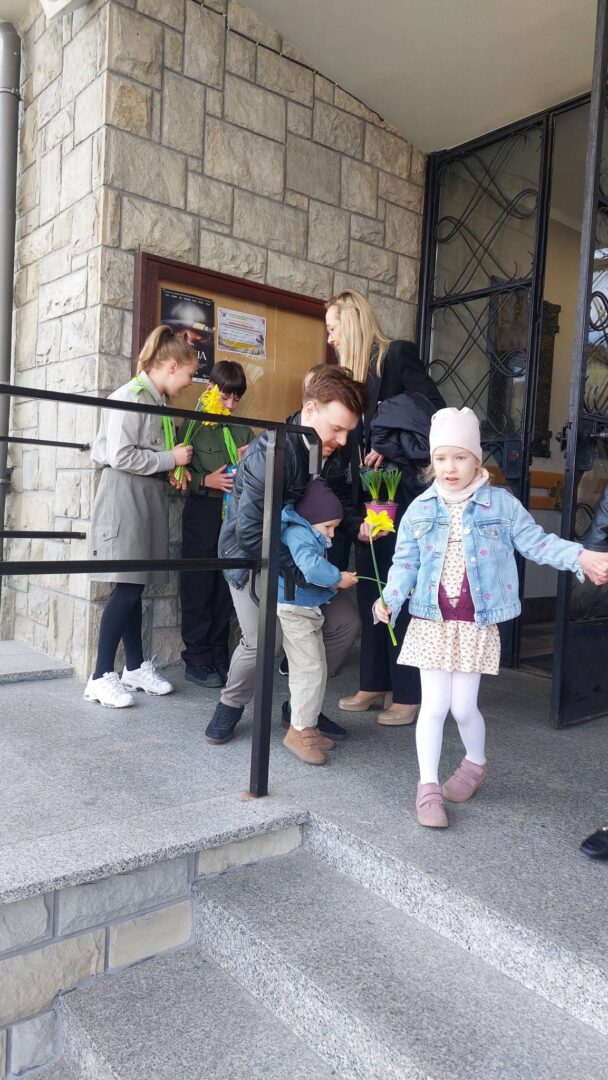 Children holding daffodils and lining up at a stone building entrance; a girl in a denim jacket stands in the foreground.