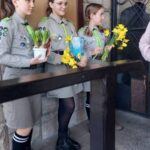 Three young Girl Scout volunteers in khaki uniforms with badges, each holding yellow daffodils outdoors by a stone wall.