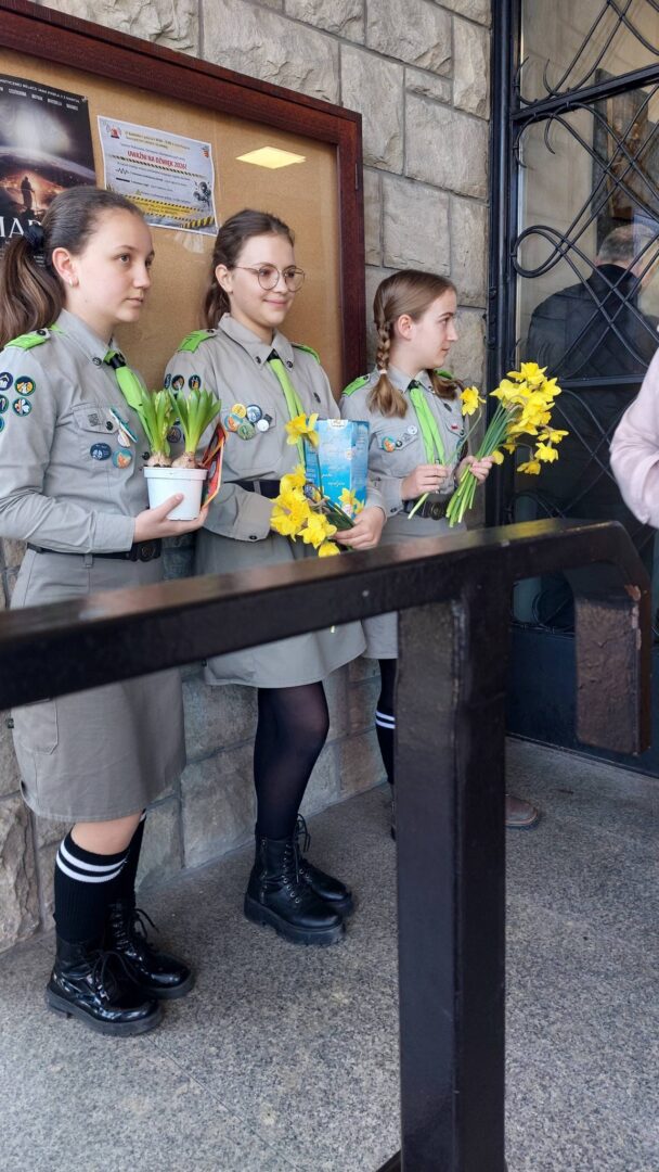 Three young Girl Scout volunteers in khaki uniforms with badges, each holding yellow daffodils outdoors by a stone wall.