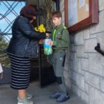 Woman in a black jacket hands a yellow daffodil bouquet and a blue gift box to a boy in a green scout uniform outside a stone building entrance.