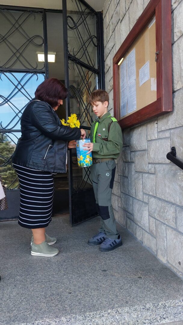 Woman in a black jacket hands a yellow daffodil bouquet and a blue gift box to a boy in a green scout uniform outside a stone building entrance.