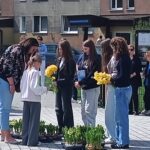 Young girl in a white hoodie receives yellow daffodils from a line of peers outdoors, with potted plants at their feet in a plaza setting nearby.