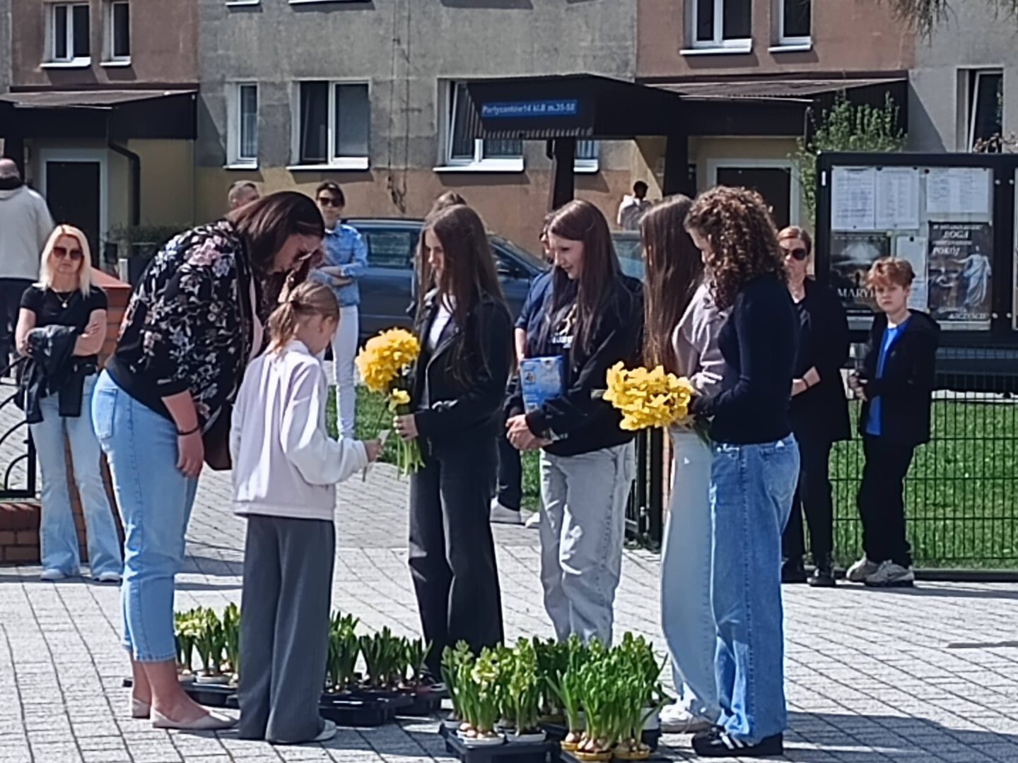 Young girl in a white hoodie receives yellow daffodils from a line of peers outdoors, with potted plants at their feet in a plaza setting nearby.